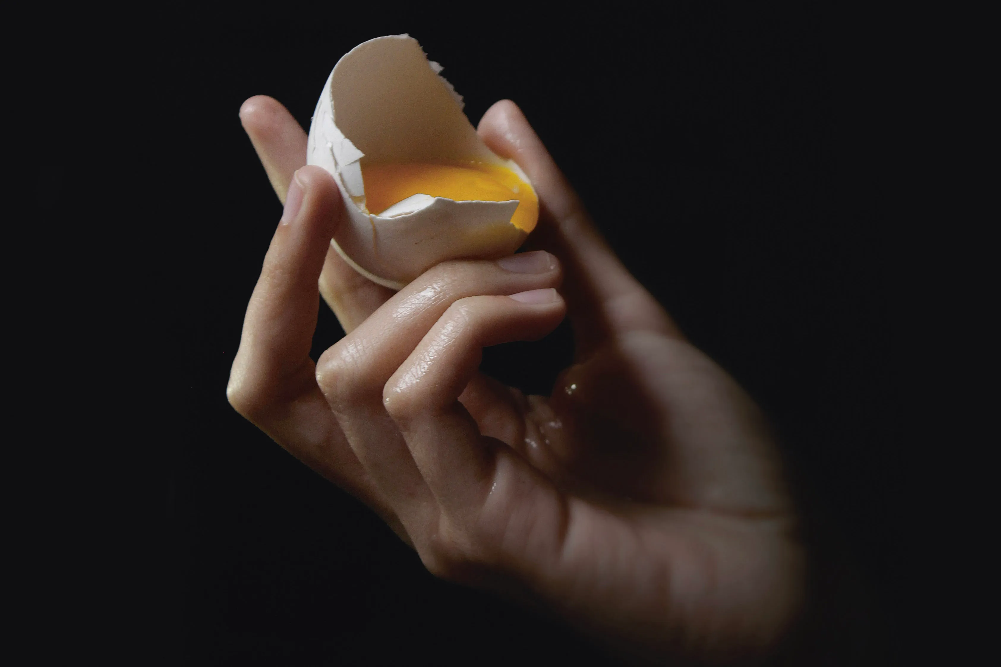 Hånd som holder en knekt eggeskall med eggeplommen synlig, mot mørk bakgrunn