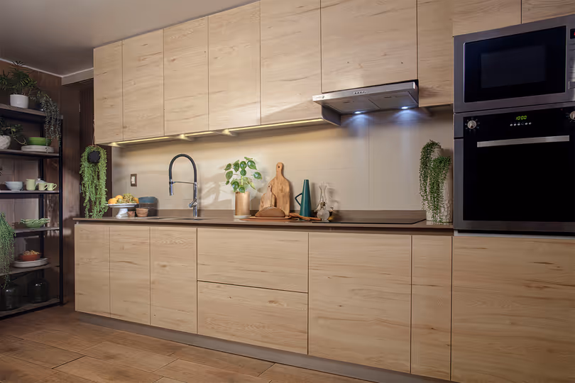 Cocina moderna con muebles de madera clara, estantes con plantas y vajilla, horno y microondas integrados, y encimera con fregadero y elementos decorativos.