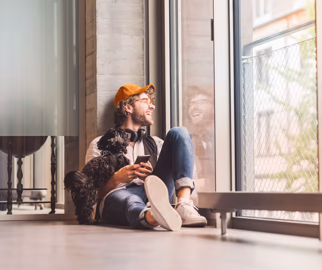 Joven sentado en el piso junto a una ventana grande, con audífonos en el cuello, sosteniendo un teléfono móvil y un perro pequeño negro apoyado en su pierna.