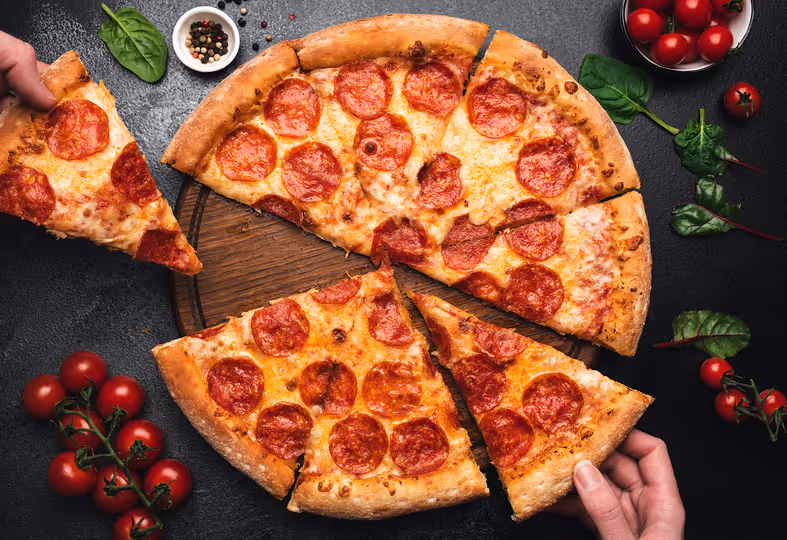 Pizza de pepperoni en tabla de madera con dos manos sosteniendo rebanadas y tomates cherry y hojas verdes alrededor.