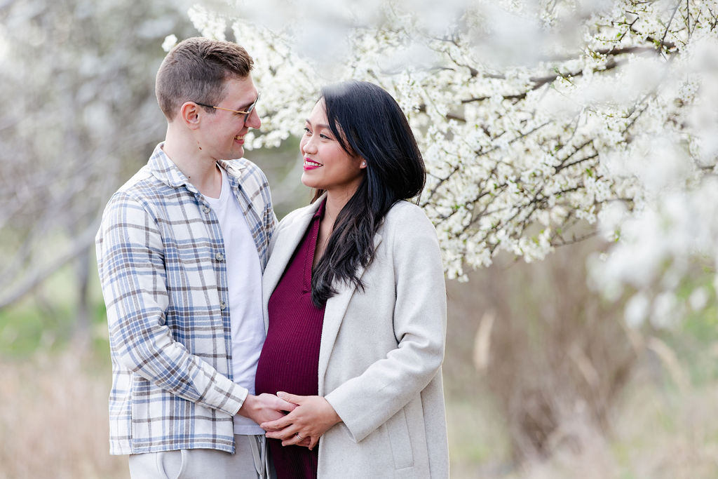 Boldog kismama és párja tavaszi kismama fotózáson egy virágzó gyümölcsösben, ami tökéletes ajándékötlet anyák napjára