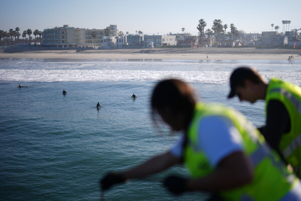 Surfers look on as Justin Hamlin, right, and Maddie Tibayan, gather a sample of seawater with a bucket off the Imperial Beach pier as part of a research team from the University of California, San Diego, Wednesday, March 11, 2026, in Imperial Beach, Calif. (AP Photo/Gregory Bull)