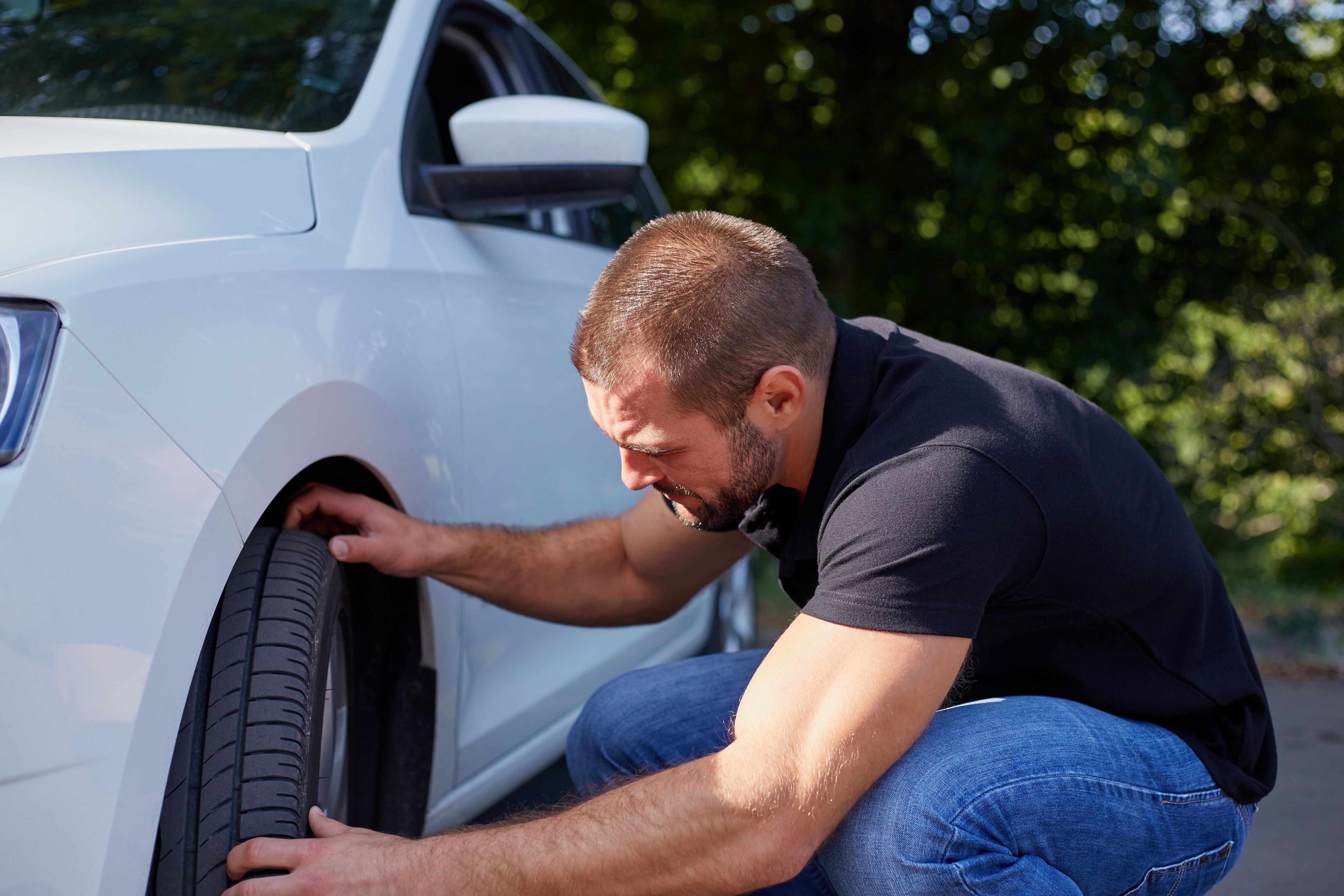Man in a black shirt and blue jeans crouching down while inspecting a car tire on a white car