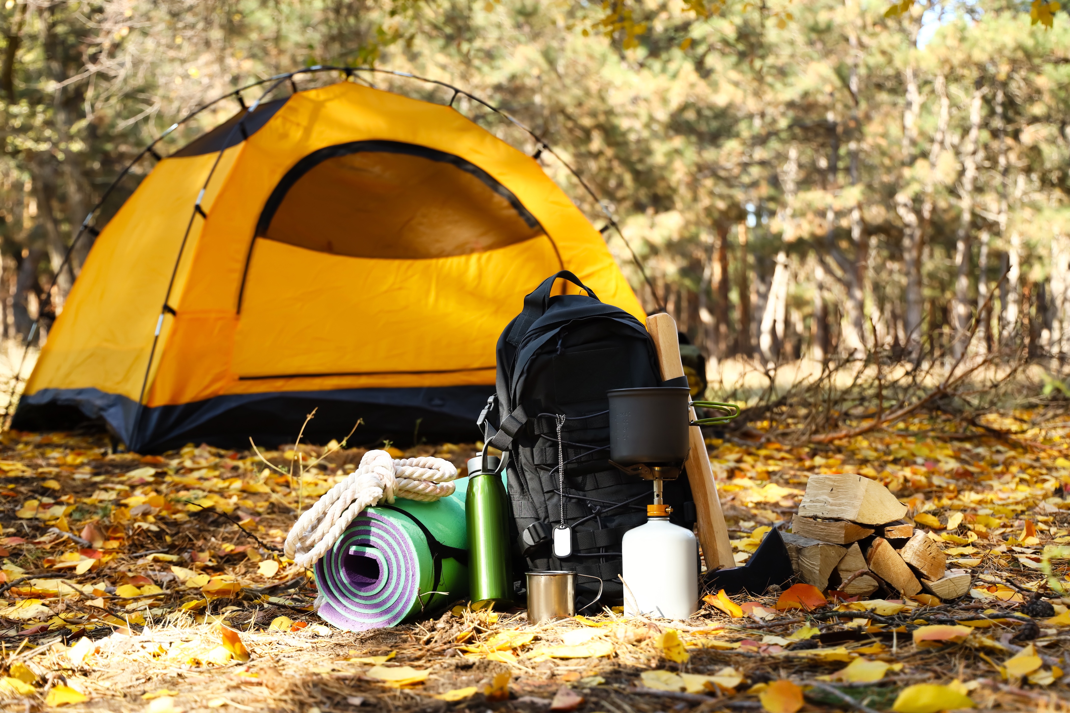 Camping backpack with camping gear sitting on the ground in front of an orange tent on a fall day in the woods
