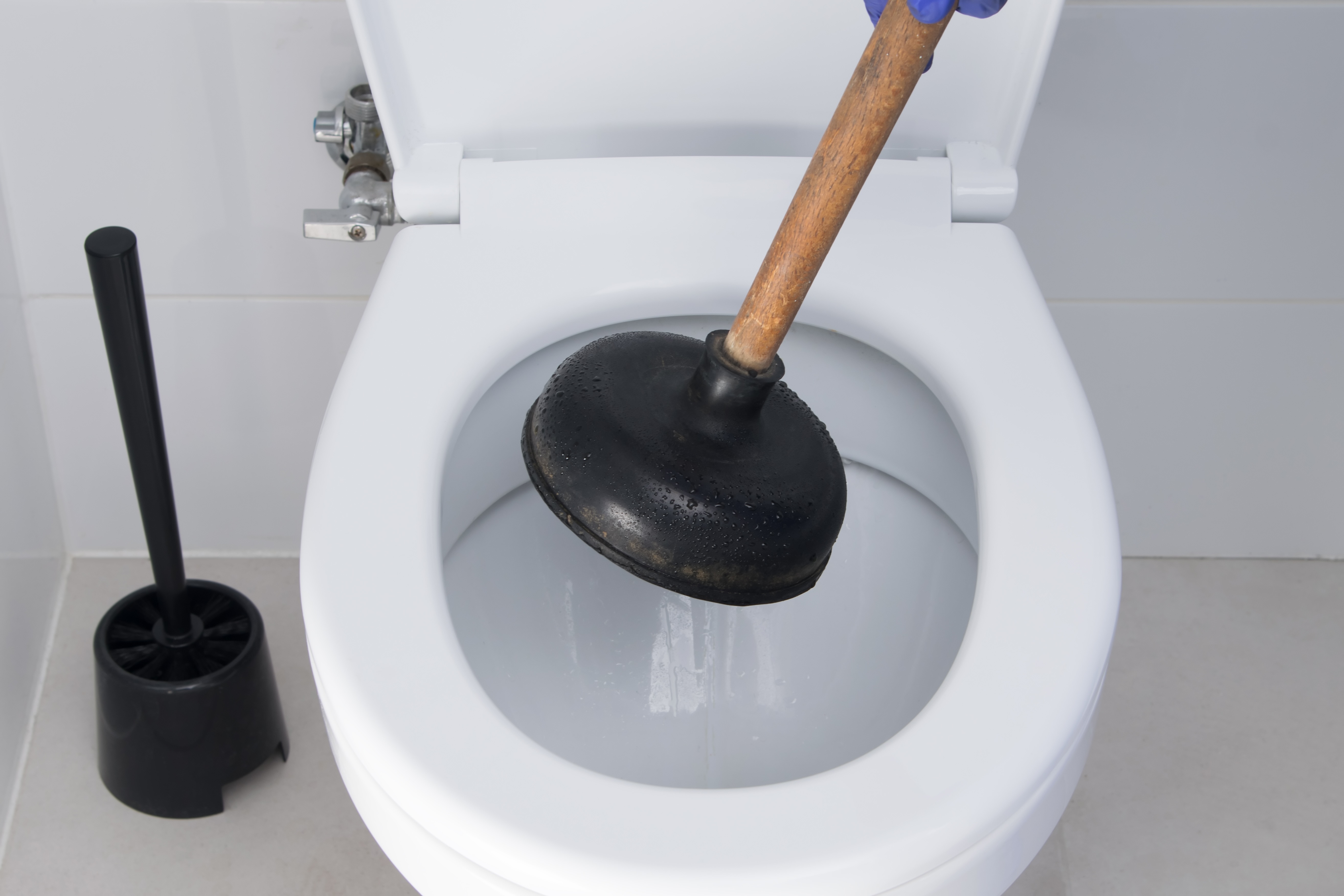 Person using a plunger to unclog a white toilet with a toilet brush in the background