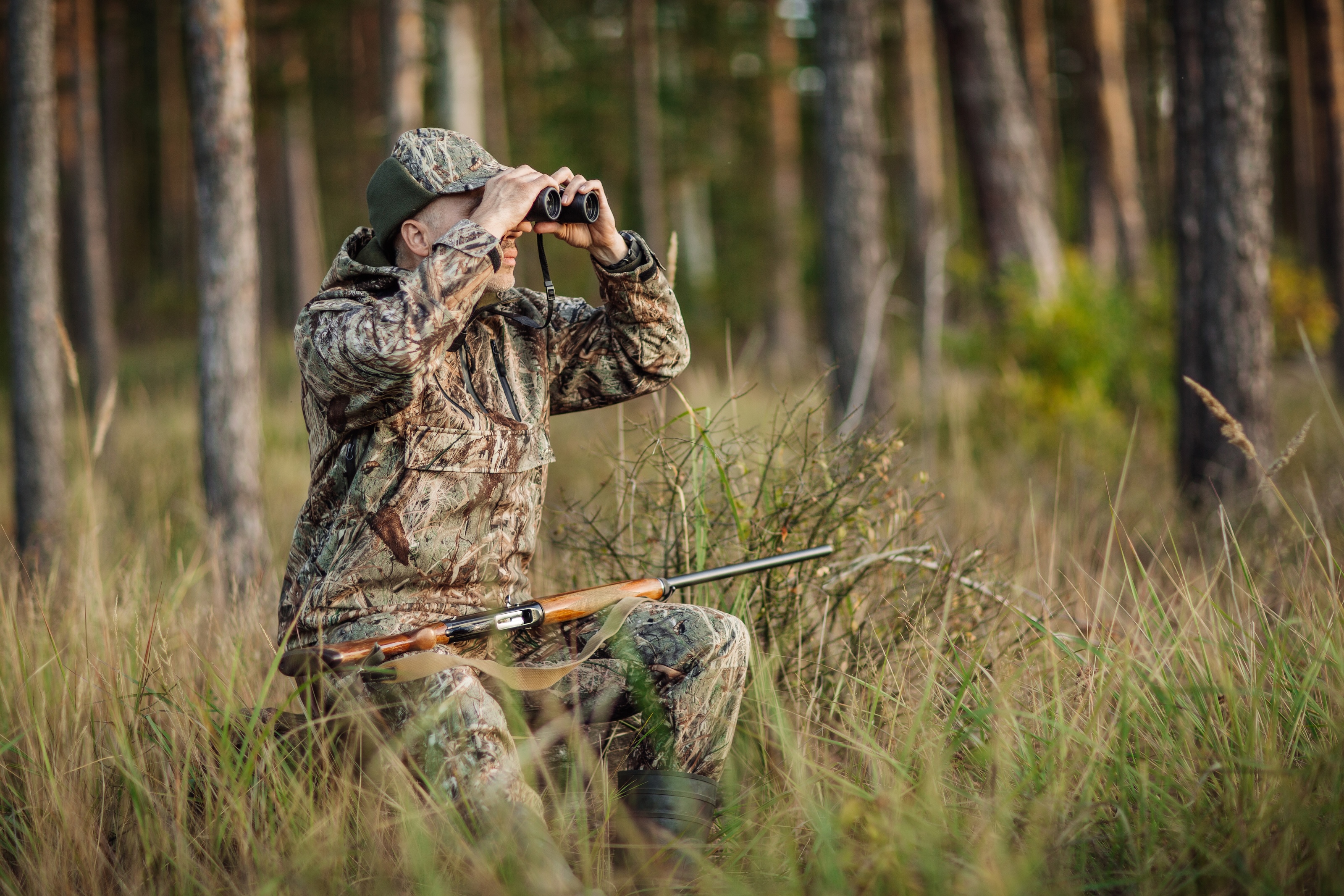 Man sitting in the woods in a full camouflage outfit and looking through binoculars
