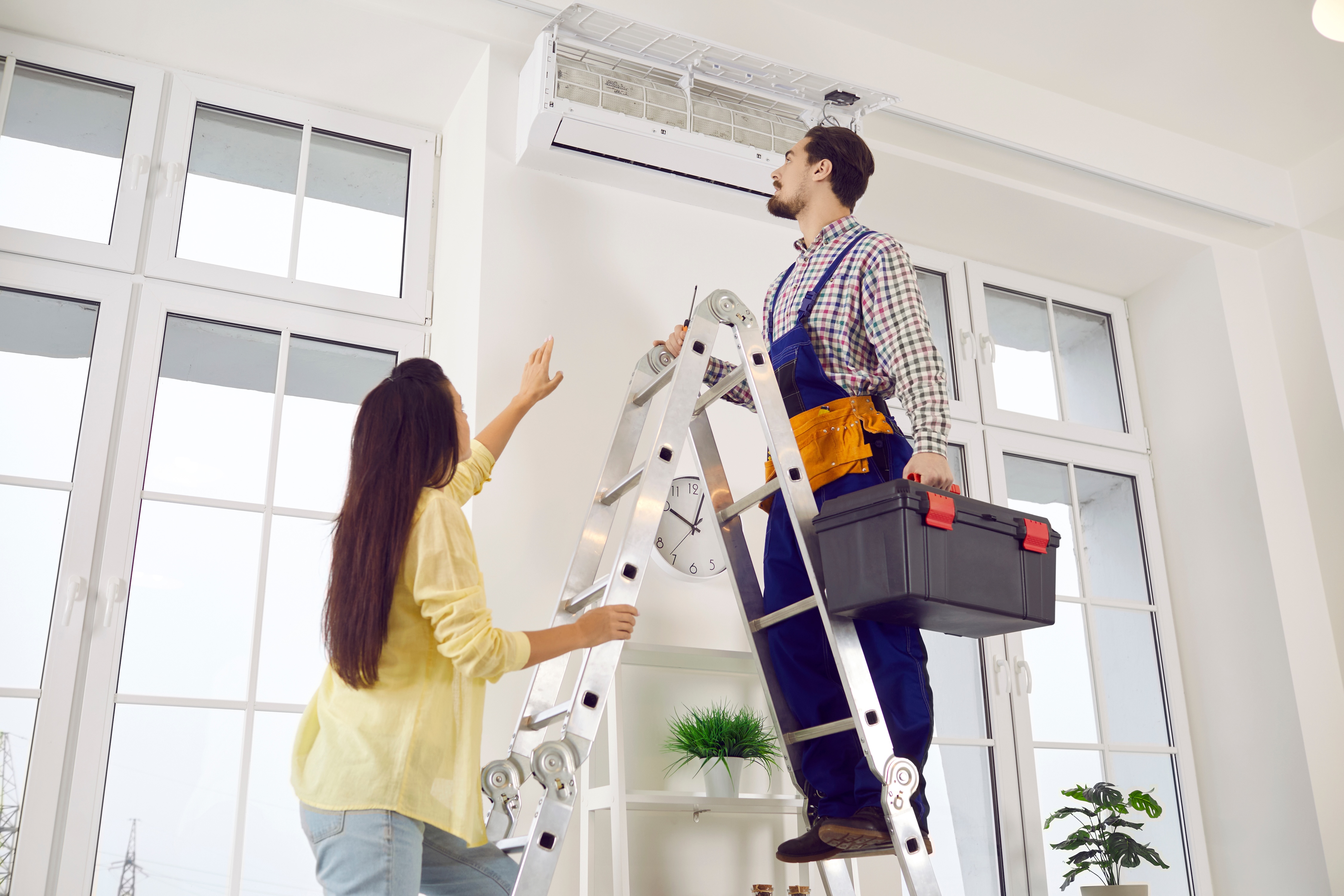 Woman talking to a repairman standing on a ladder to look at an AC unit