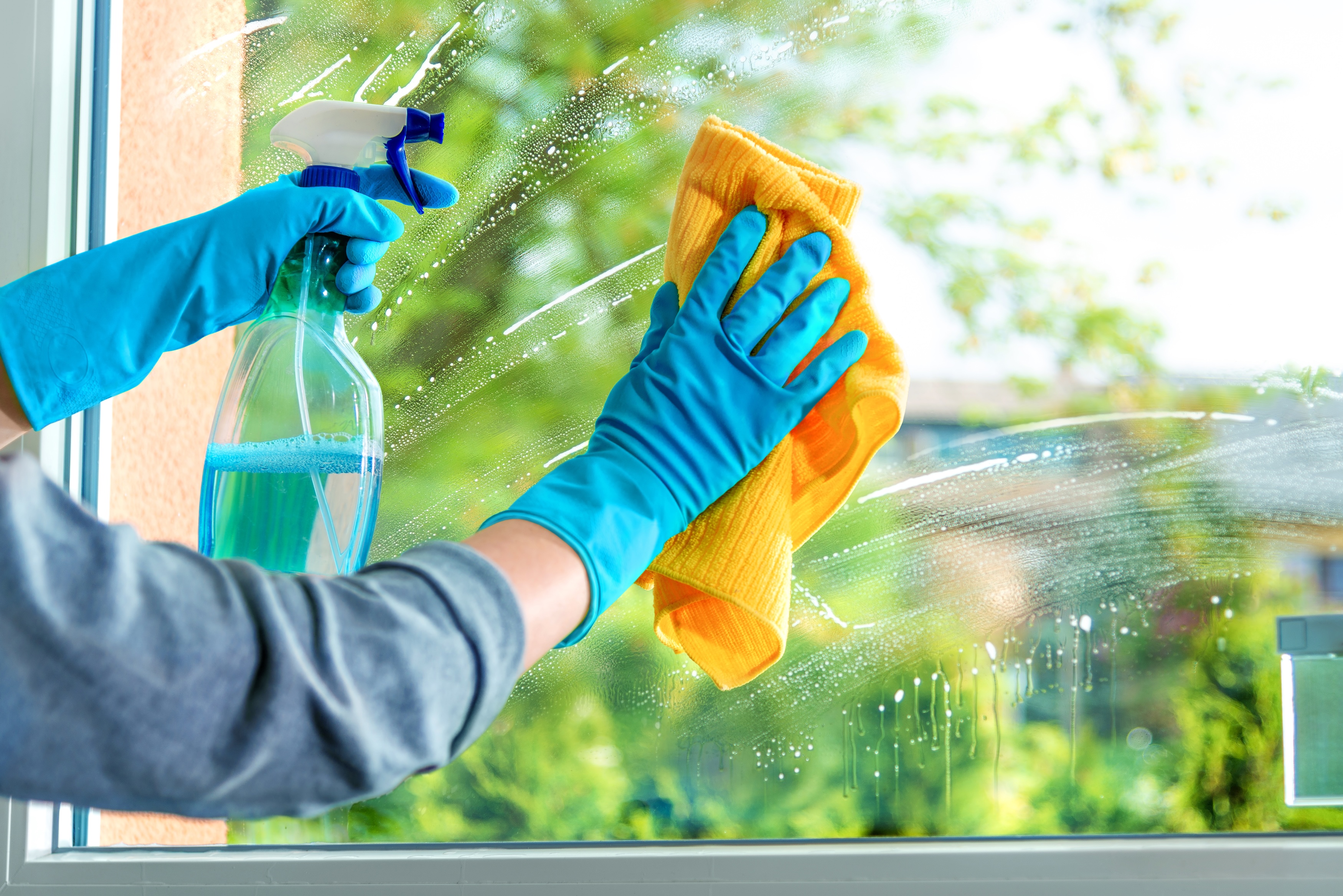 Person wearing blue gloves, cleaning a window with a yellow cloth and a spray bottle