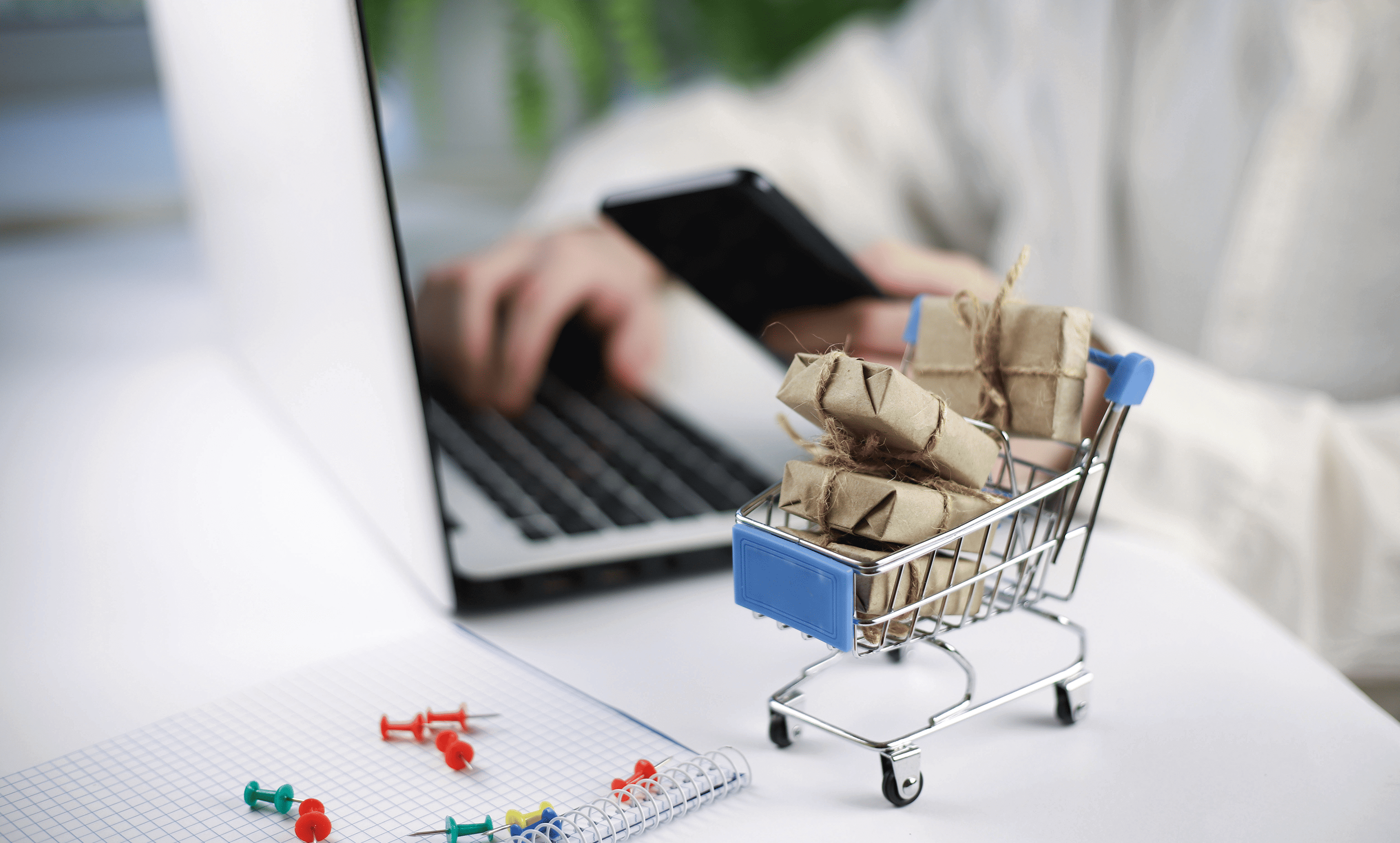 Small shopping cart filled with cardboard boxes on desk next to laptop, representing online shopping and e-commerce.