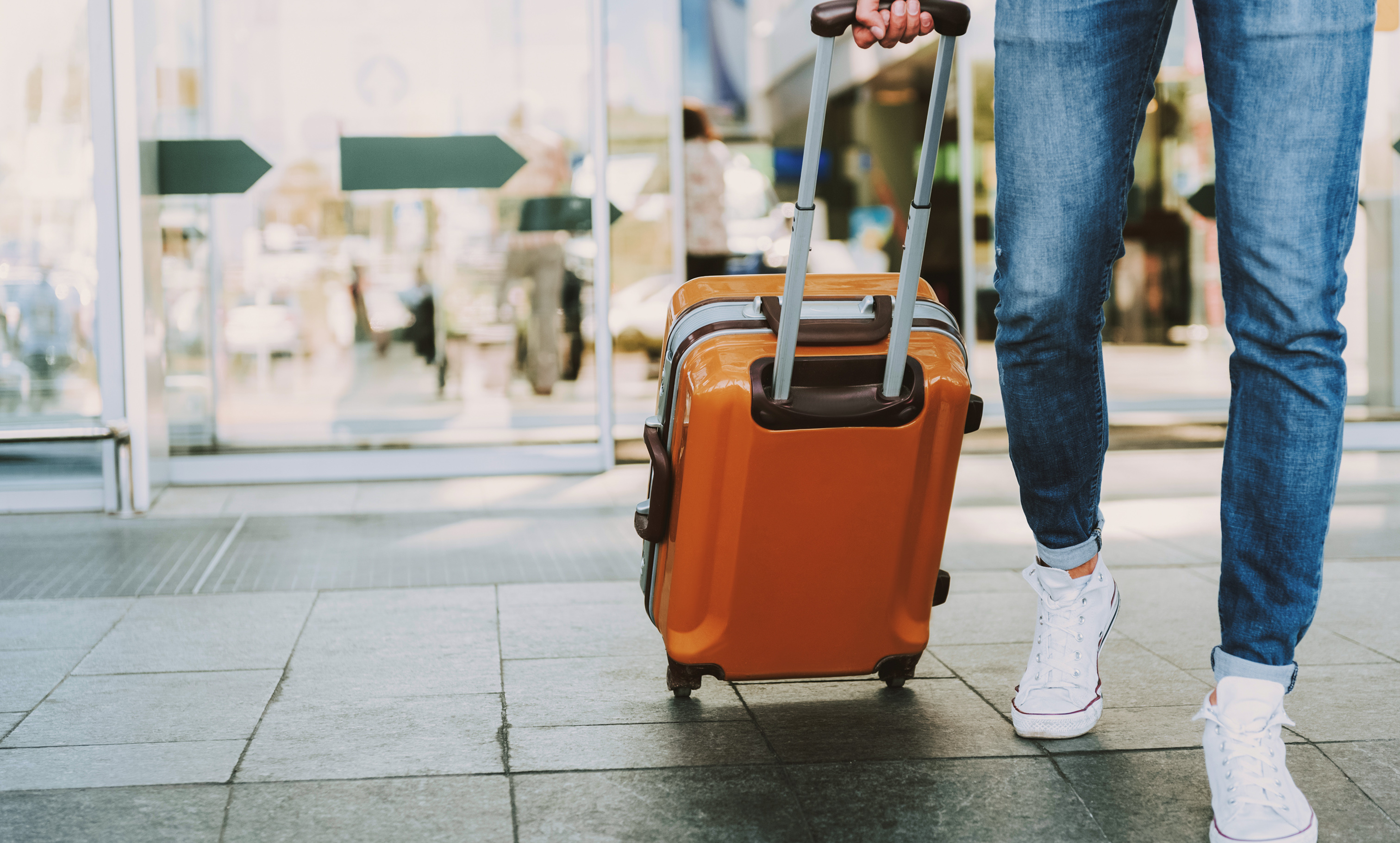 Person in jeans and white sneakers pulling an orange rolling suitcase through an airport terminal with directional signs.