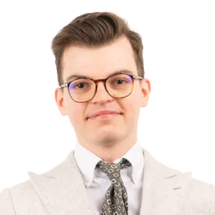 Professional headshot of a man wearing glasses, beige blazer, white shirt, and patterned tie against white background.