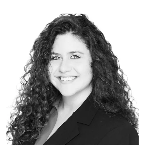 Professional headshot of a woman with long curly dark hair, wearing a black blazer, smiling at the camera against a white background.