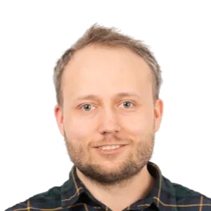 Headshot of a smiling man with light brown hair and beard wearing a dark checkered shirt against a white background.