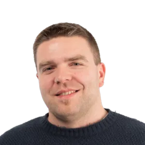 Headshot of a smiling man with short brown hair wearing a dark sweater against a white background.