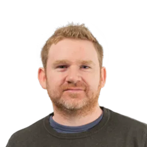 Headshot of a man with light brown hair and facial hair wearing a dark colored shirt against a white background.