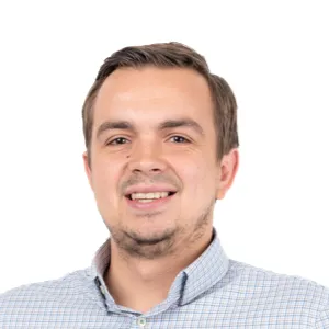 Professional headshot of a smiling man with brown hair wearing a light blue checkered shirt against a white background.