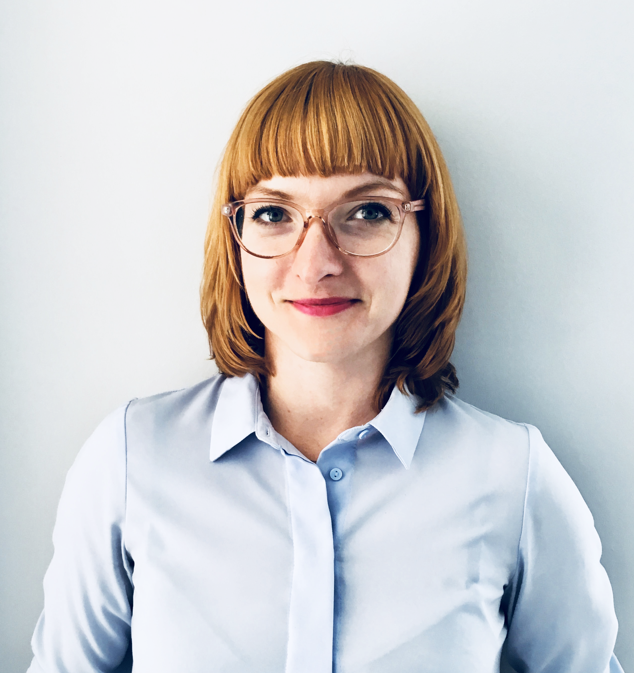 Professional headshot of a woman with reddish-brown hair and bangs, wearing glasses and a light blue button-up shirt against a white background.