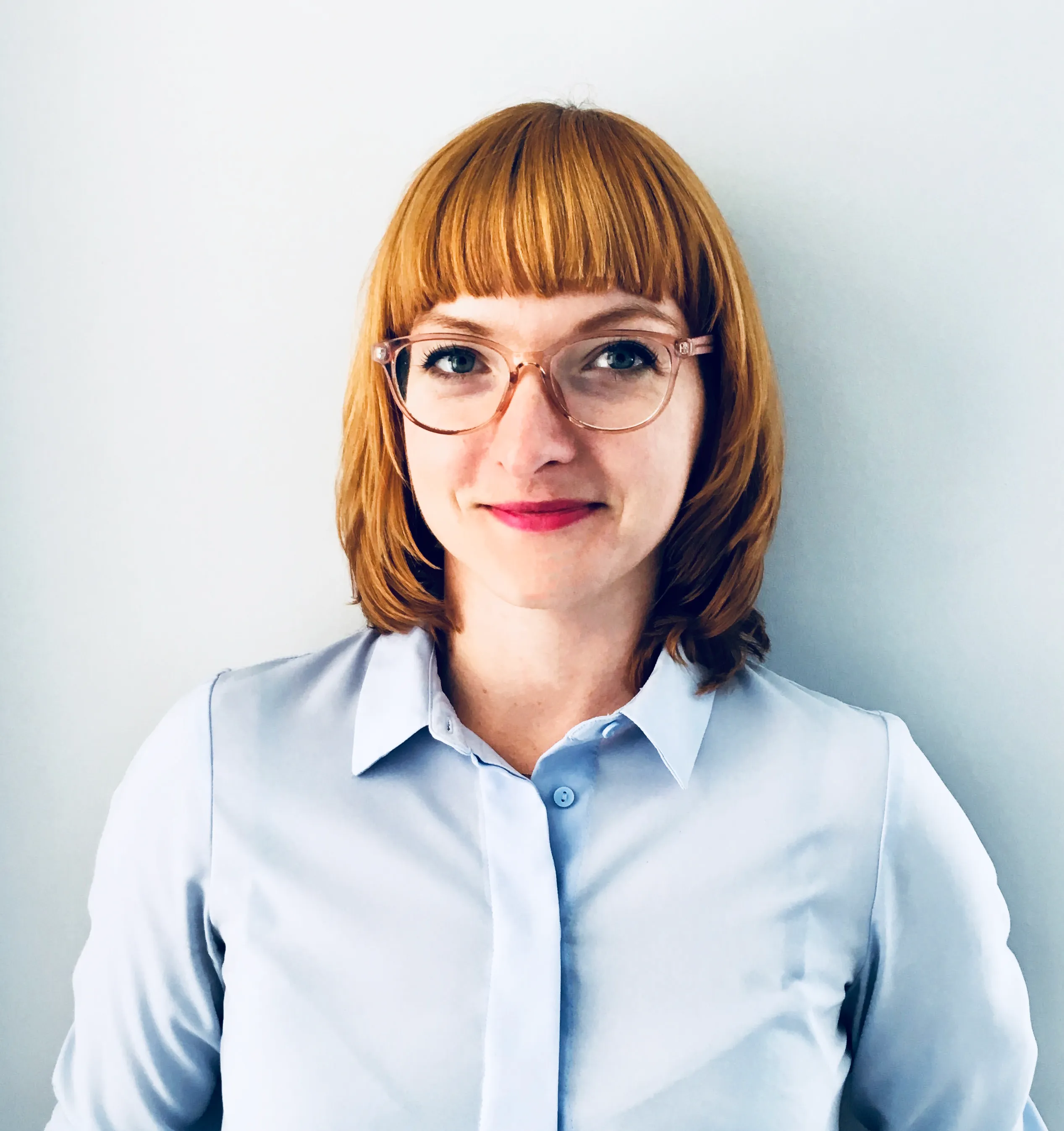 Professional headshot of a woman with reddish-brown hair and bangs, wearing glasses and a light blue button-up shirt against a white background.