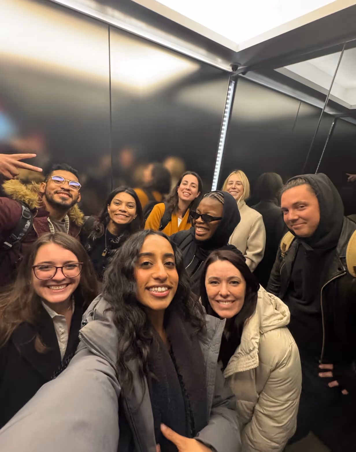 Group of nine smiling friends taking a selfie inside an elevator with mirrored black walls.