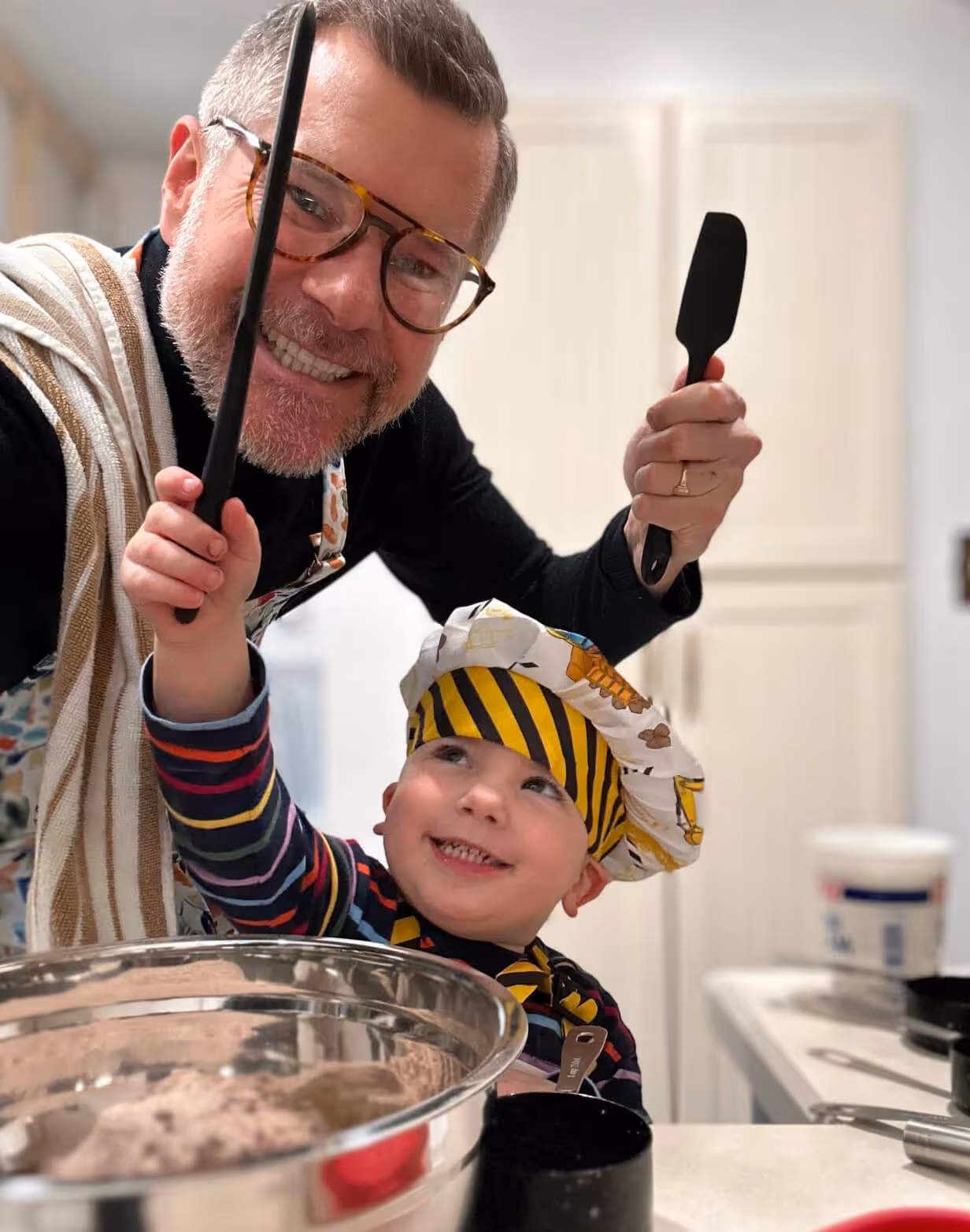 Smiling man and young child in kitchen playing with spatulas, child wearing a chef's hat and striped shirt.