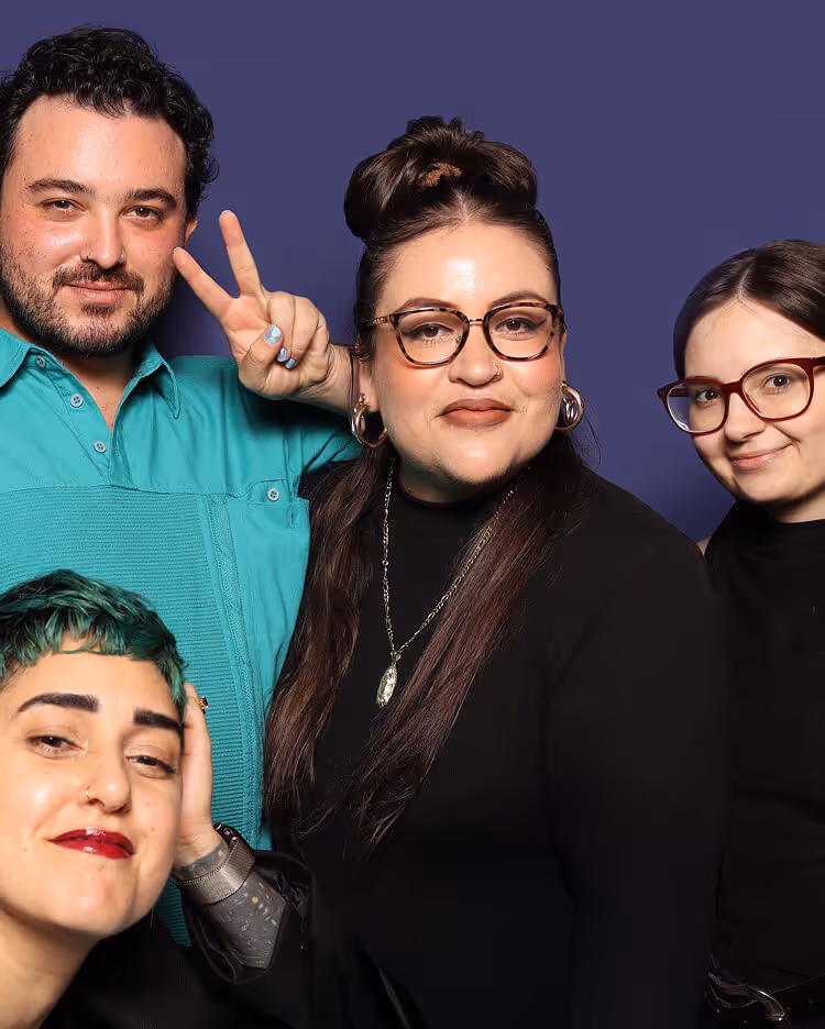 Group portrait of four smiling young adults against a dark blue background, one man in teal shirt making a peace sign.