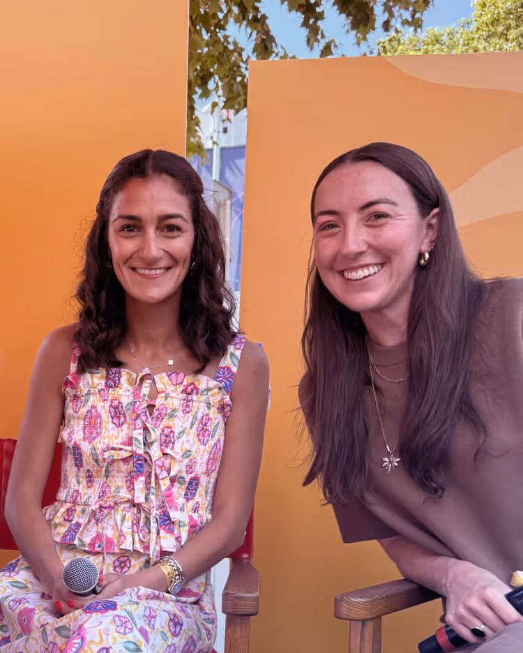 Two smiling women sitting in chairs with yellow backgrounds, one wearing a floral dress holding a microphone and the other in a brown shirt holding a microphone.
