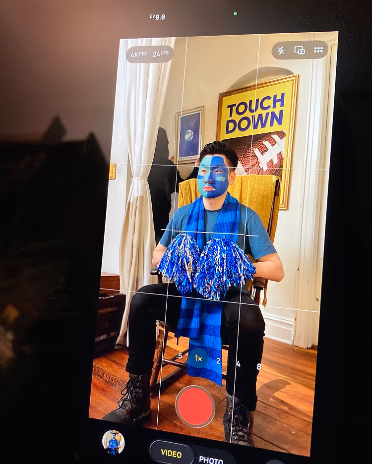 Person with blue face paint sitting on a yellow chair, holding blue pom-poms, in front of a 'TOUCH DOWN' football poster, captured through a smartphone camera interface.