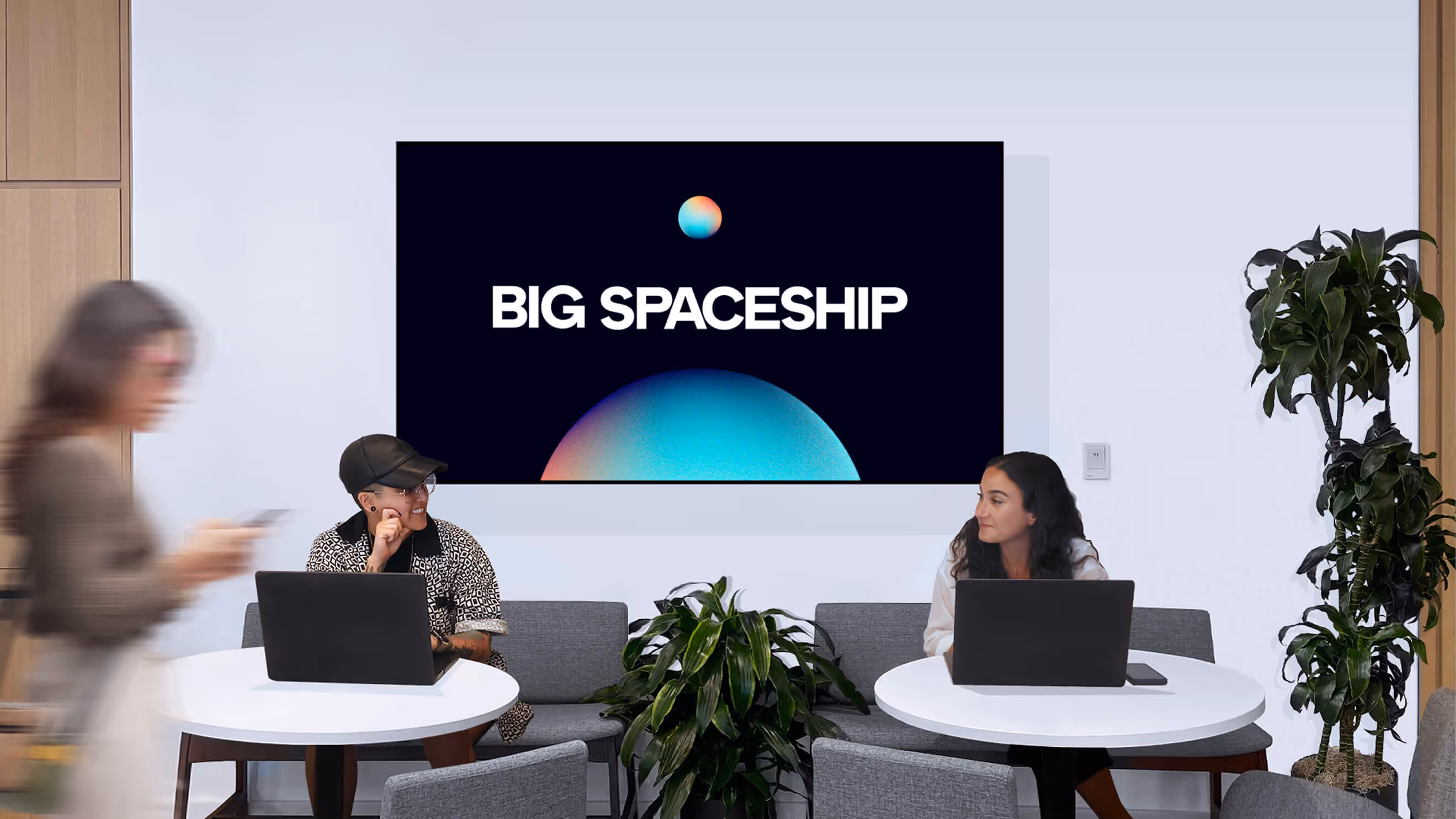 Two people sitting at separate round tables with laptops, facing each other, under a wall display reading 'BIG SPACESHIP' with a blue and pink planet graphic.