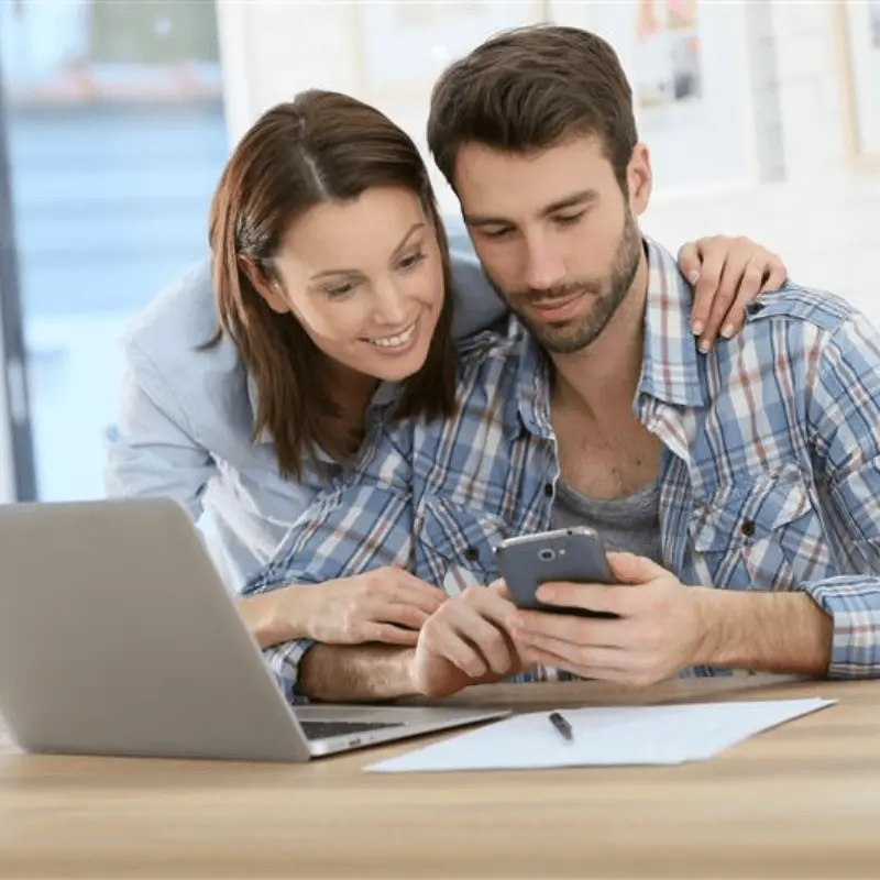 Happy couple reviewing information on phone and laptop, getting guidance through trusted housing counseling services.