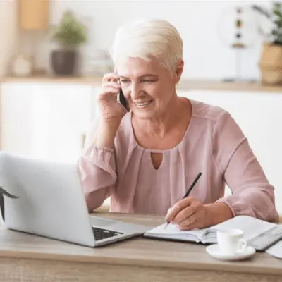 A woman smiling while seated at a table with a laptop and pen, participating in reverse mortgage counseling.