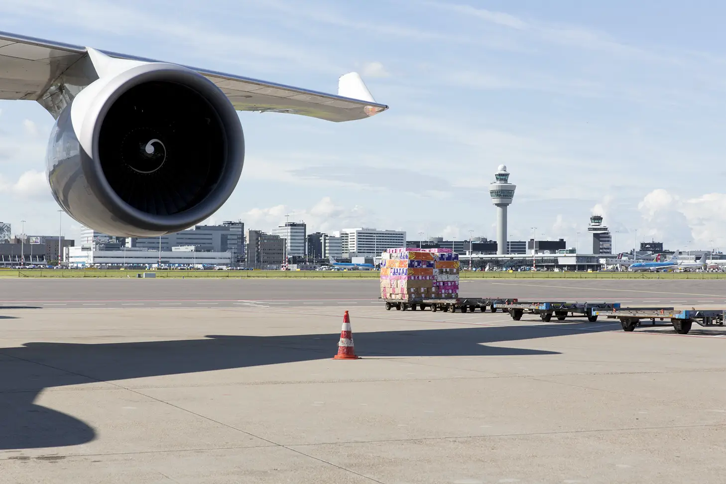 Close-up of an airplane engine at an airport with cargo loaded on containers and control towers in the background under a cloudy sky.