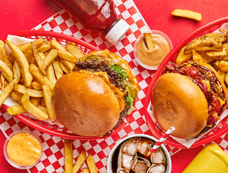 Deux burgers servis avec des frites, différentes sauces, une bouteille de ketchup et un verre de soda avec une paille rouge et blanche sur une table rouge.
