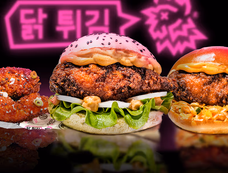 Deux burgers au poulet frit avec de la laitue, des oignons et une sauce, accompagnés de deux morceaux de poulet frit épicé, sur fond noir avec un panneau lumineux rose en coréen.