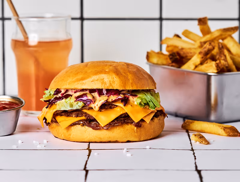 Burger au cheddar avec laitue, sauce et chou rouge, accompagné de frites et d'un verre de boisson.