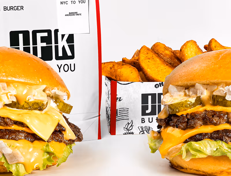Deux cheeseburgers avec fromage, laitue, cornichons, oignons et sauce, accompagnés de quartiers de pommes de terre frits dans un emballage blanc imprimé.