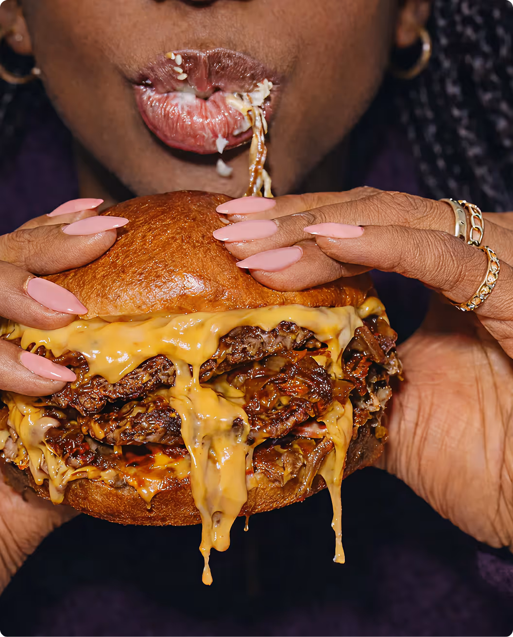 Person with polished pink nails holding a dripping cheeseburger, biting into it with melted cheese stretching from their mouth.