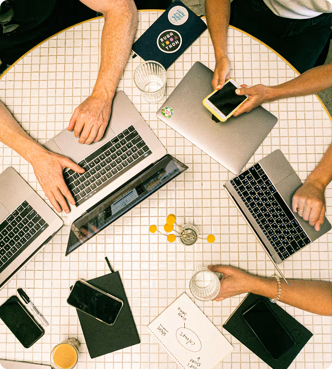 Vue de dessus d'une table ronde avec plusieurs ordinateurs portables, téléphones, verres d'eau et carnets, avec des personnes interagissant autour.