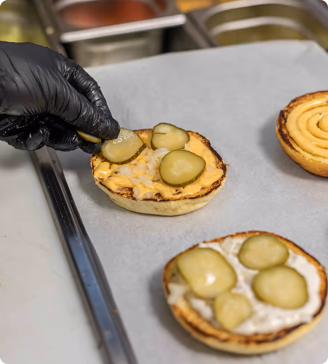 Une main gantée noire place des tranches de cornichon sur un pain à hamburger avec sauce et oignons hachés.
