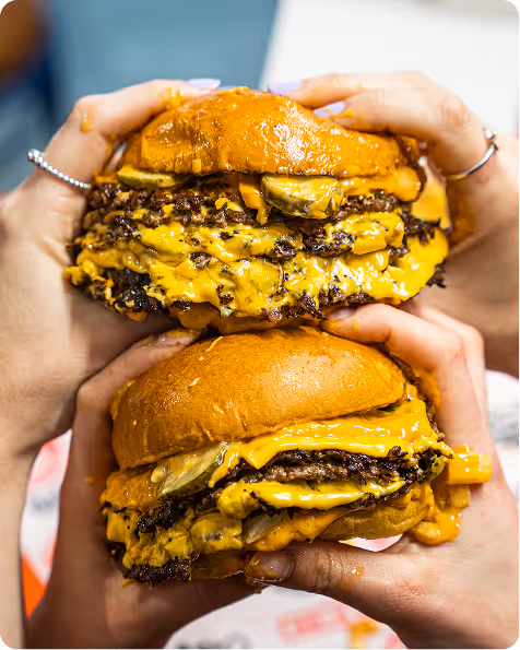 Deux mains tenant deux gros burgers cheeseburgers fondus avec plusieurs tranches de fromage et cornichons.