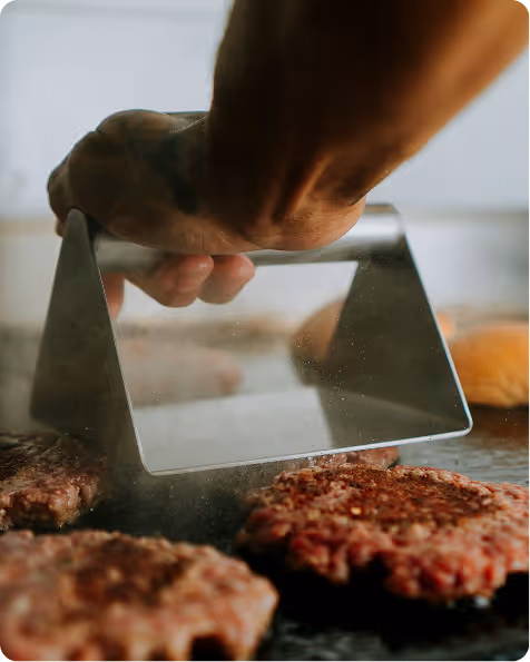 Une main utilisant une presse pour aplatir des steaks hachés crus sur une plaque de cuisson.