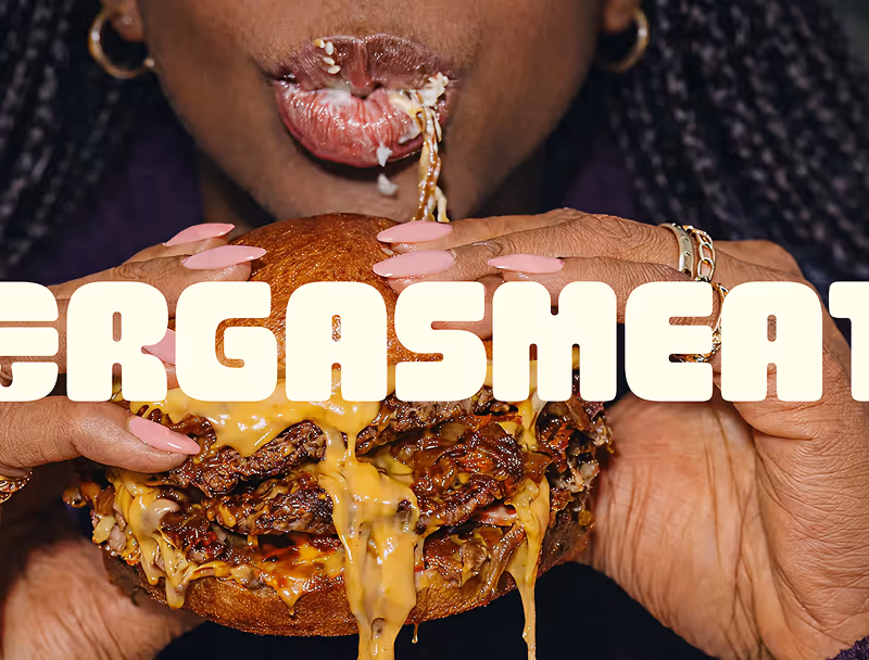 Une personne tenant un hamburger double avec beaucoup de fromage fondu et mastiquant une bouchée, avec le texte partiel 'ERGASMEAT' superposé.