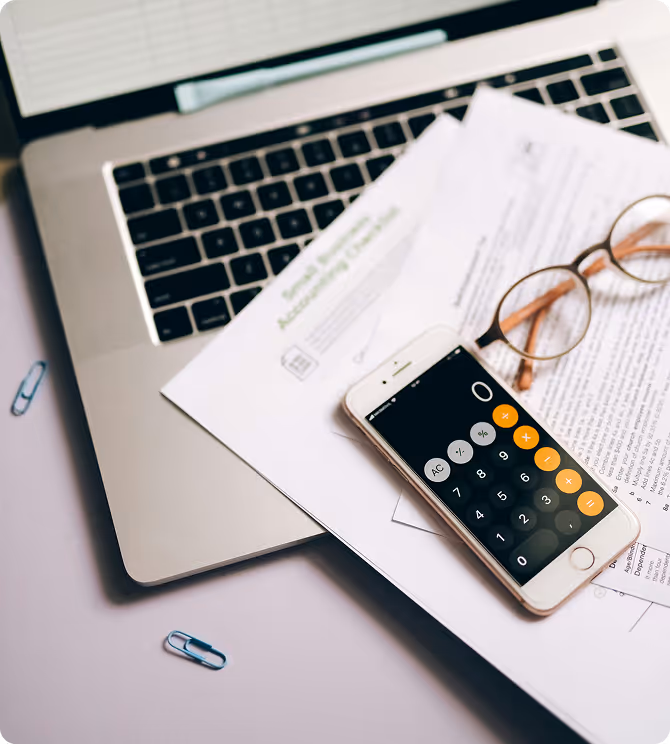 Ordinateur portable avec des documents, une paire de lunettes et un smartphone affichant une application calculatrice posés dessus.