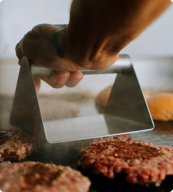 Main tenant une presse à hamburger en acier au-dessus de steaks hachés sur un gril.
