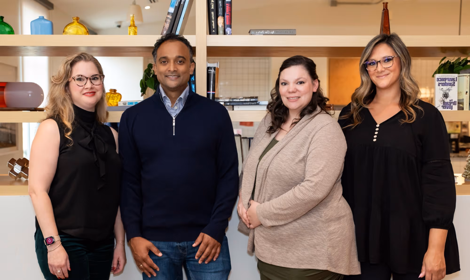 Group of two women and one man standing and smiling in front of a wooden bookshelf with decorative items and books.