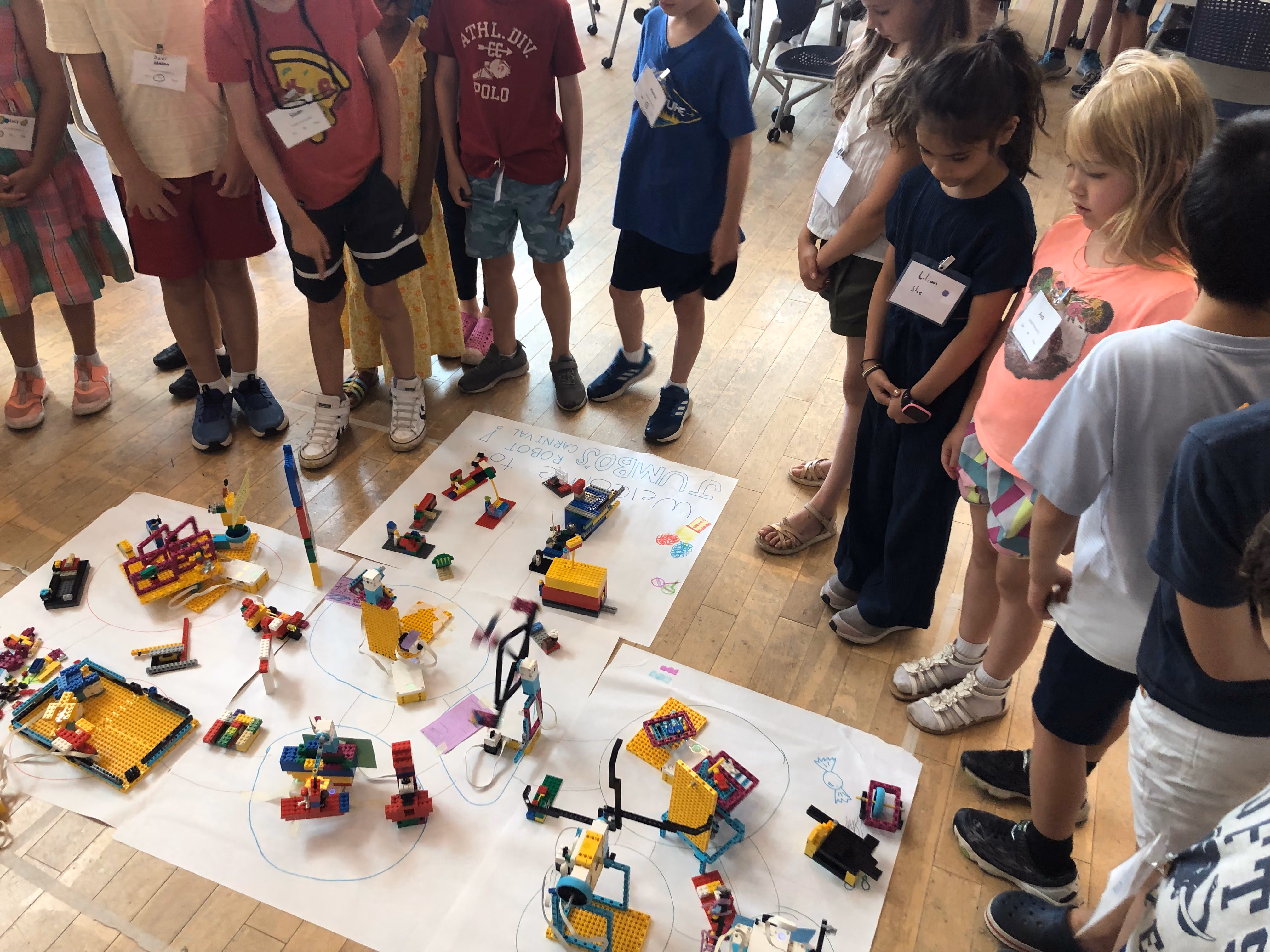 Group of children with name tags standing around a bunch of LEGO robots on large pieces of paper on the floor