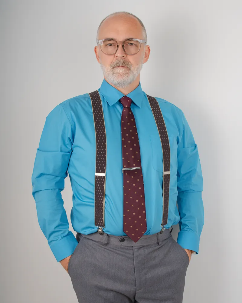 Studio portrait of a man wearing glasses, a blue shirt, tie and suspenders.