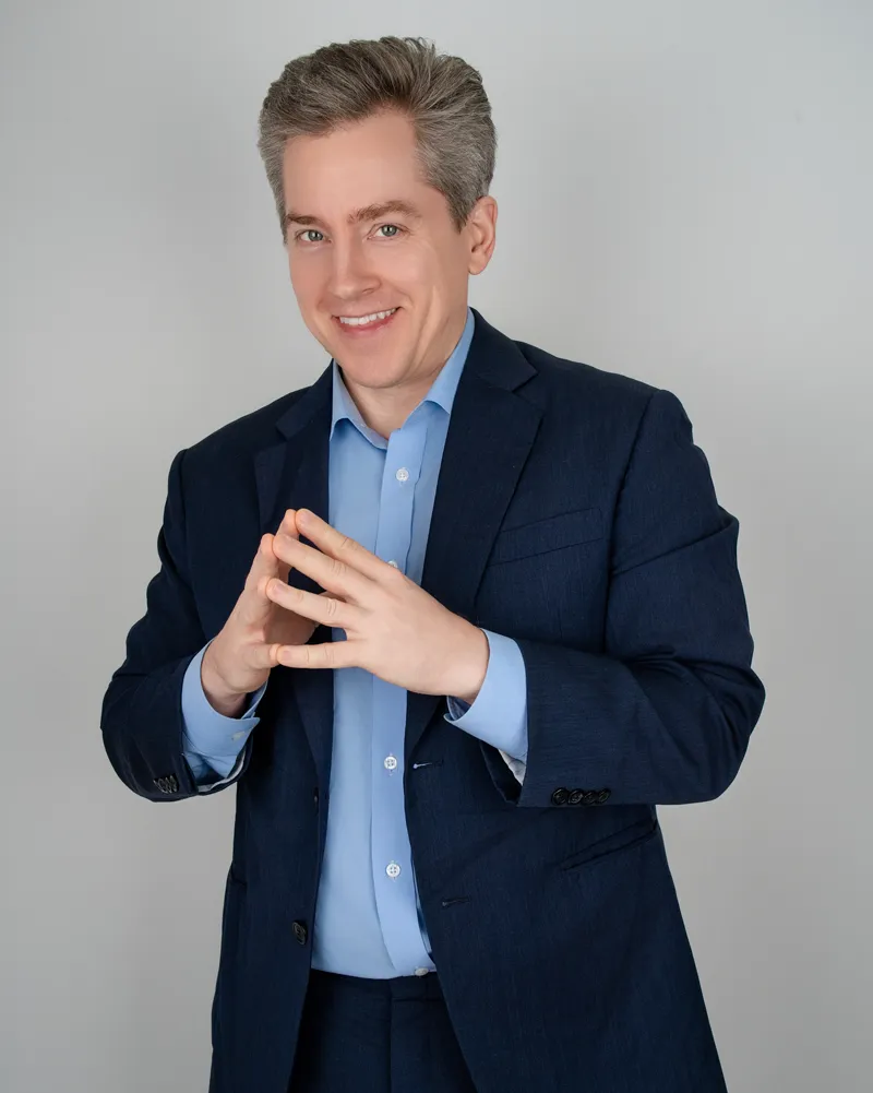 Studio portrait of a smiling man in a navy suit with his hands together.
