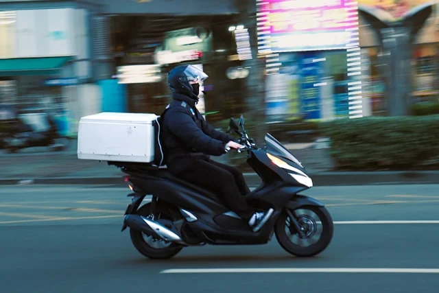 A delivery rider carrying a white delivery box on a motor scooter.