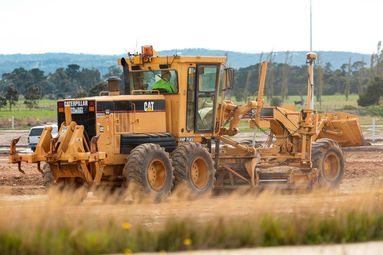 Yellow Caterpillar motor grader levels dirt road at construction site, operator in cab, large tires kicking up dust in daylight.