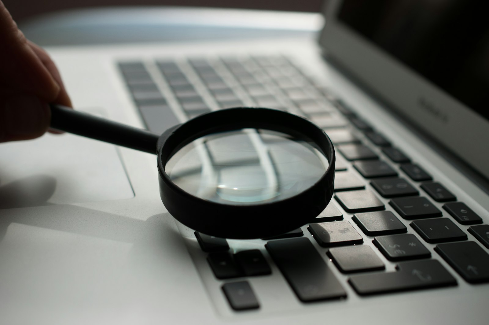 A hand holds a magnifying glass over a laptop keyboard, inspecting it closely, illustrating software inspection and testing.